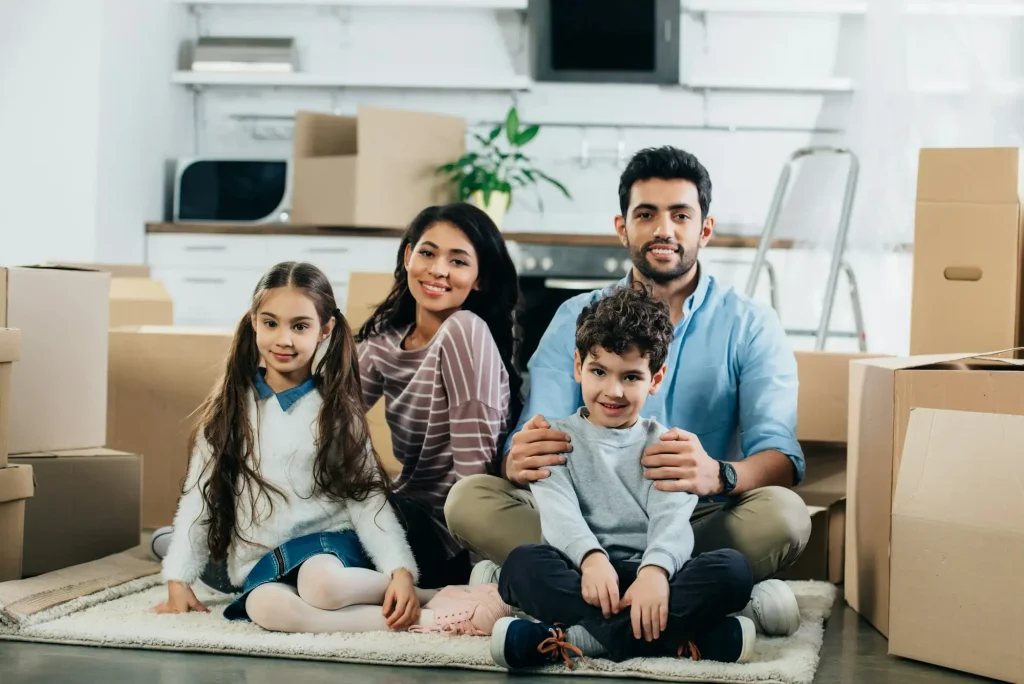 happy latin husband and wife sitting in carpet with cute children in new home