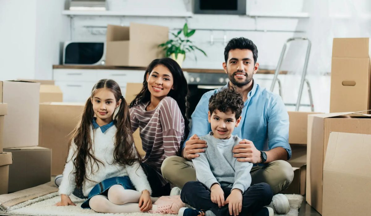 happy latin husband and wife sitting in carpet with cute children in new home
